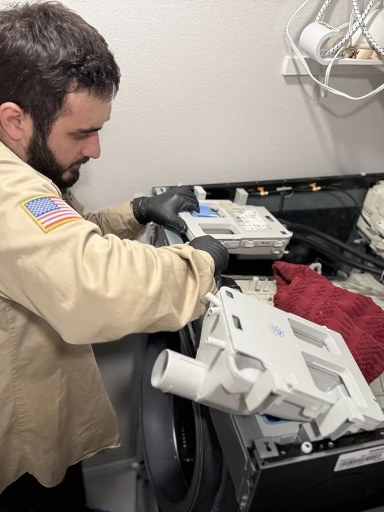 Alpha and Omega technician holding the detergent dispenser assembly removed from an open front-load washer